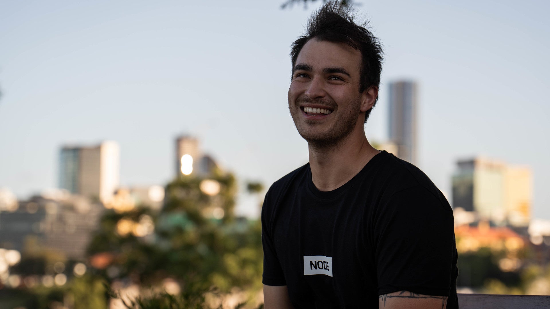 A man in a black NODE t-shirt smiling, city skyline behind him.