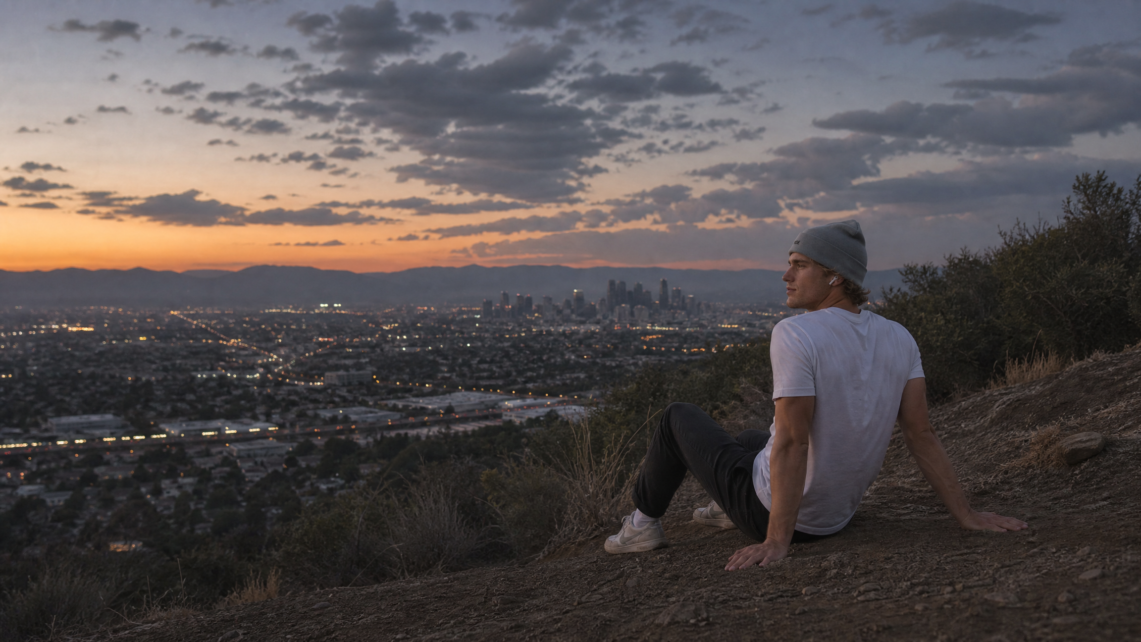 A person in a NODE hoodie sitting on a hillside above a city at sunset.